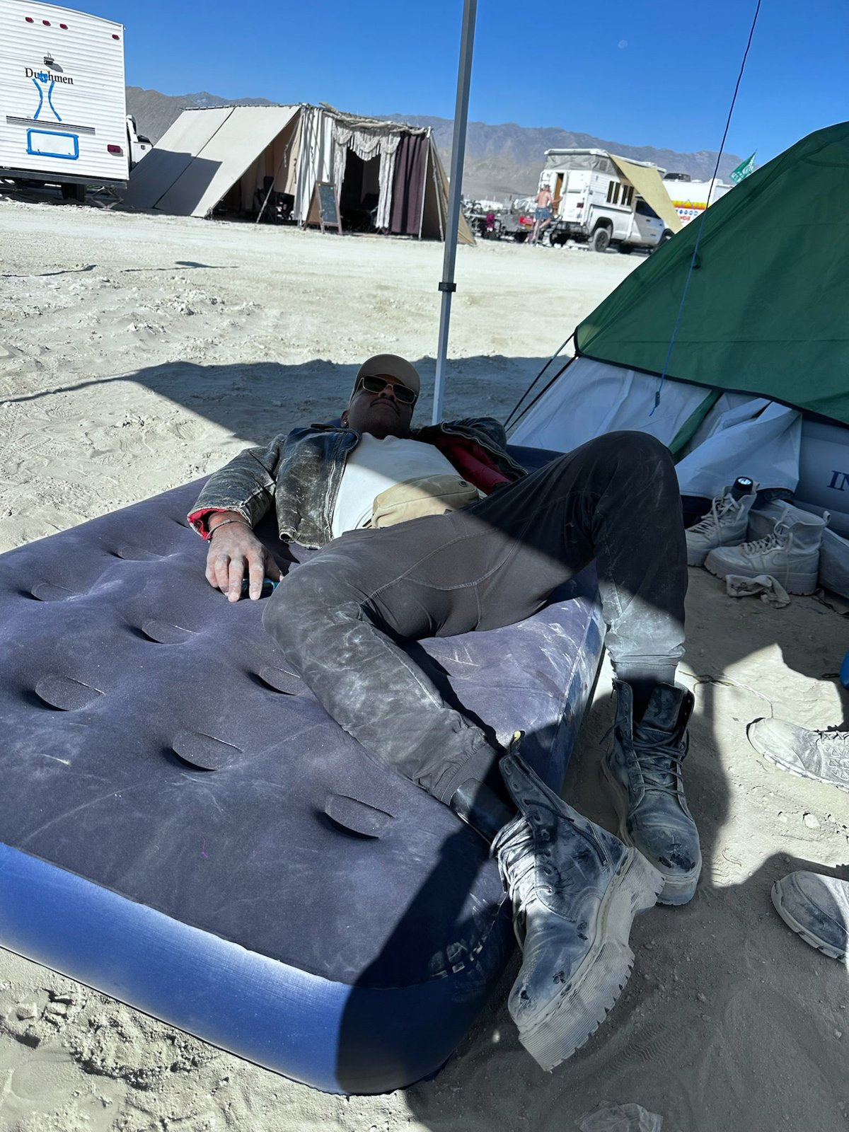 Camper resting on an air mattress in the shade next to a tent at a dusty desert festival camp